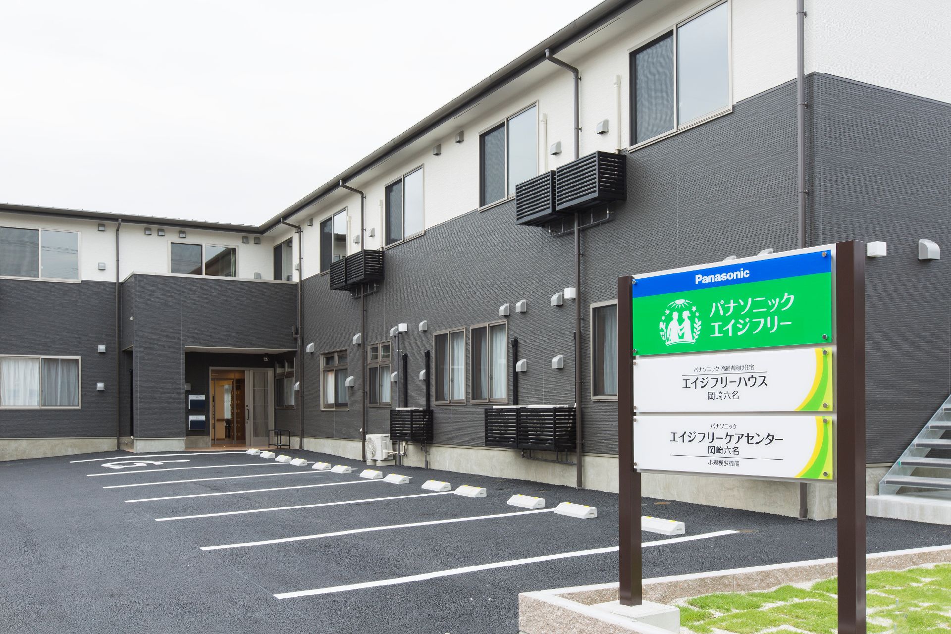 白とグレーの2階建ての建物。玄関先には看板があり、屋外階段もある。