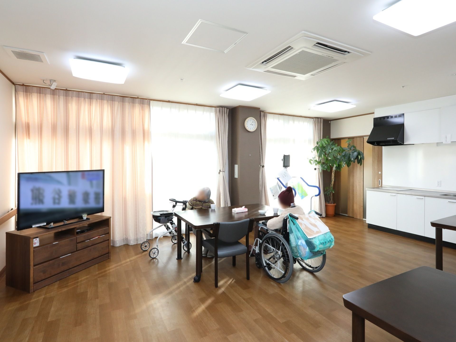 フローリングの床に、ダイニングテーブルが置かれている。テレビが設置されている。大きな窓があり、レースのカーテンが掛かっている。