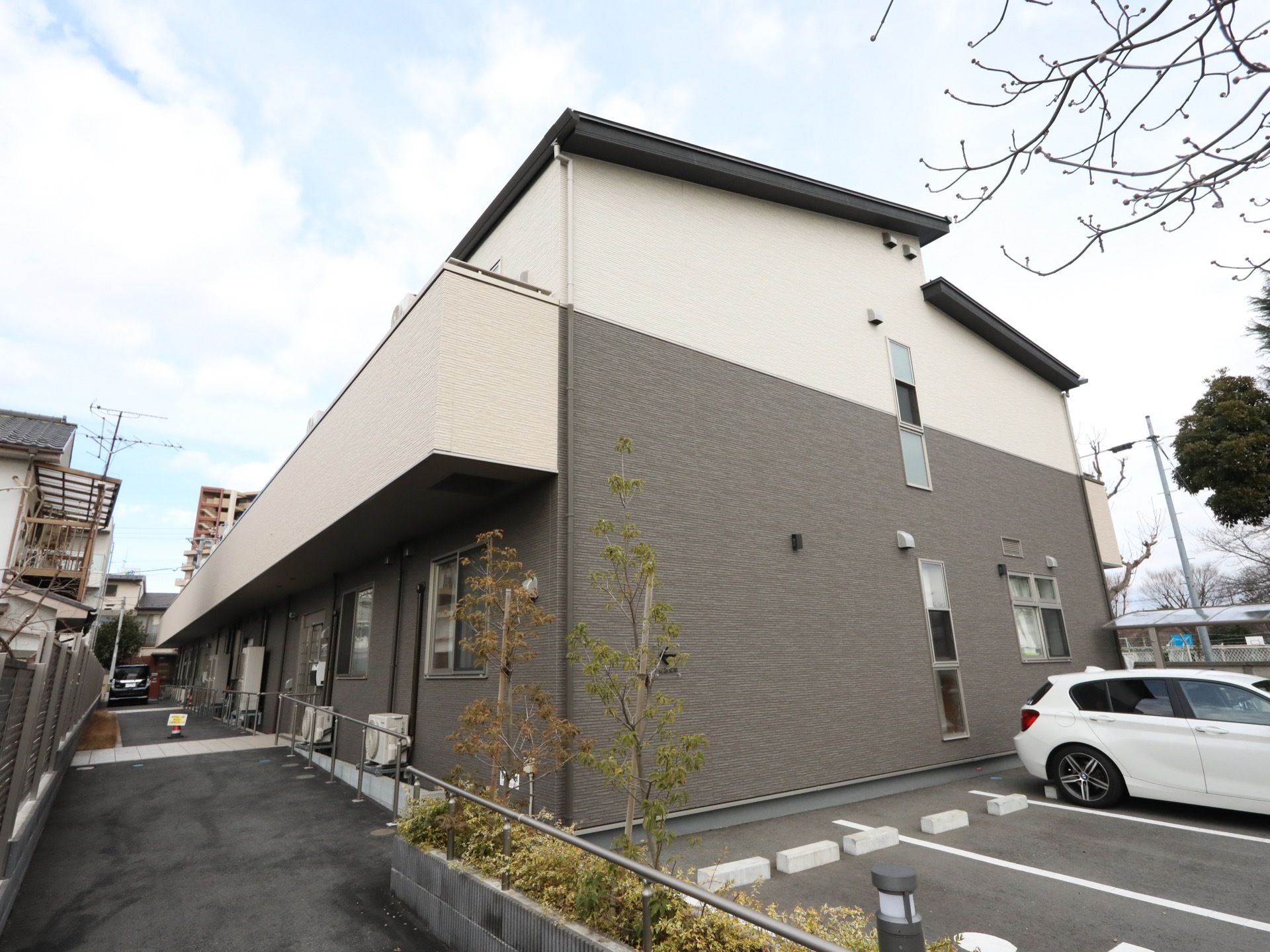 茶色と白のタイルが貼られた２階建て住宅が建っている。敷地は舗装されており、駐車場には車が１台停められている。