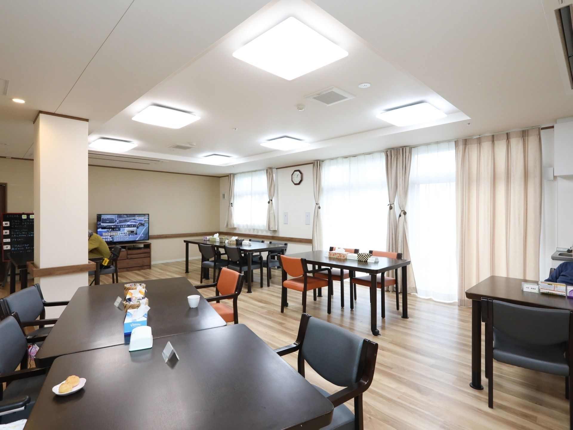 広々としたフロアにテーブルセットが並べられている。奥にテレビが設置され、入居者様が１人くつろいでいる。