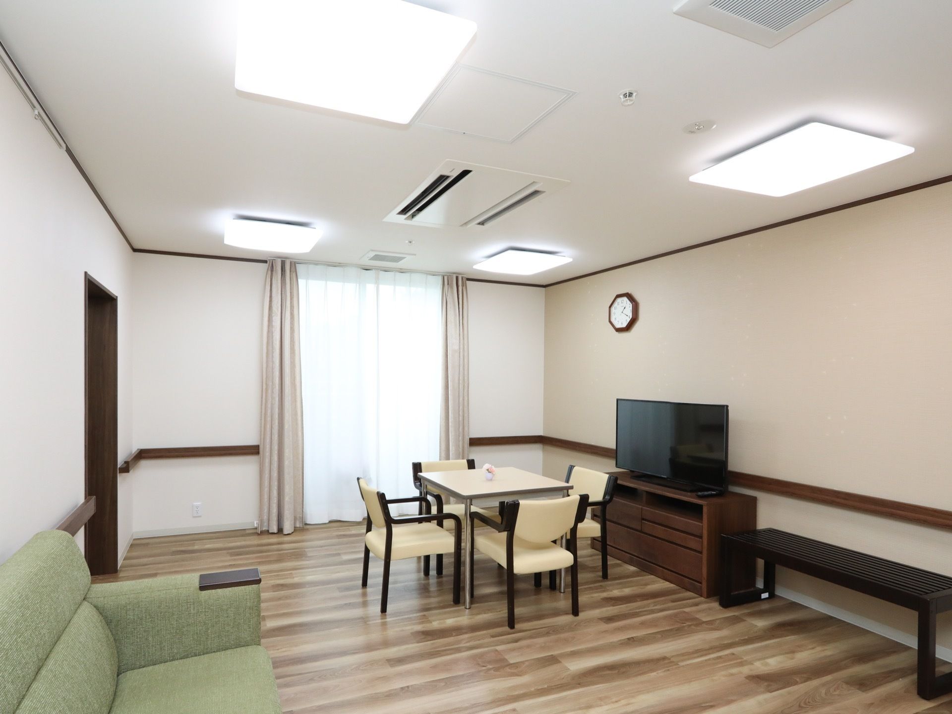掃き出し窓から陽の入っている明るいスペースがある。天井には空調があり、テレビやテーブルセットが置かれている。