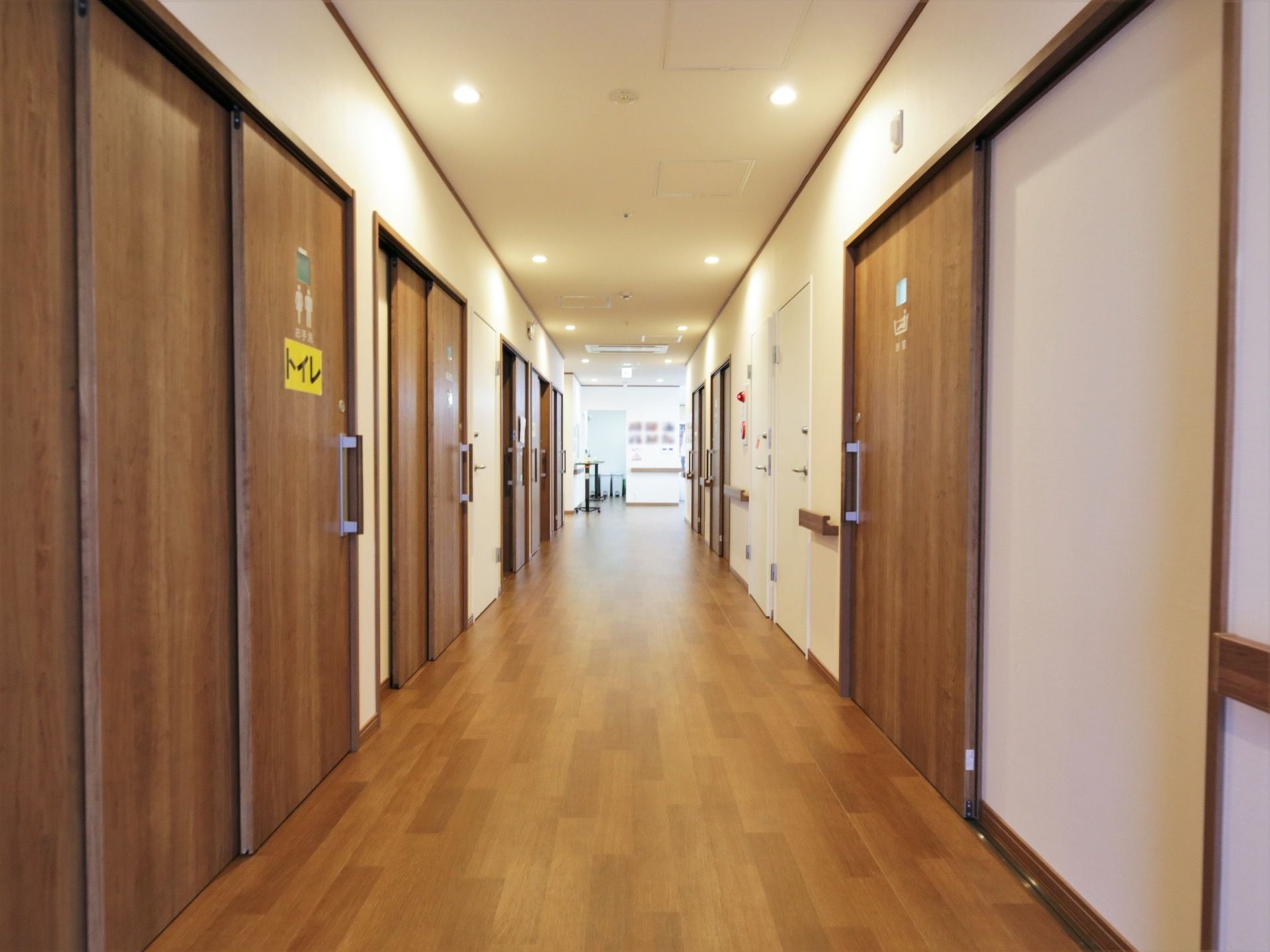 白い壁と木目調の床とのコントラストがおしゃれな通路である。共用トイレの扉には、わかりやすい大きな文字でトイレの表示がされている。