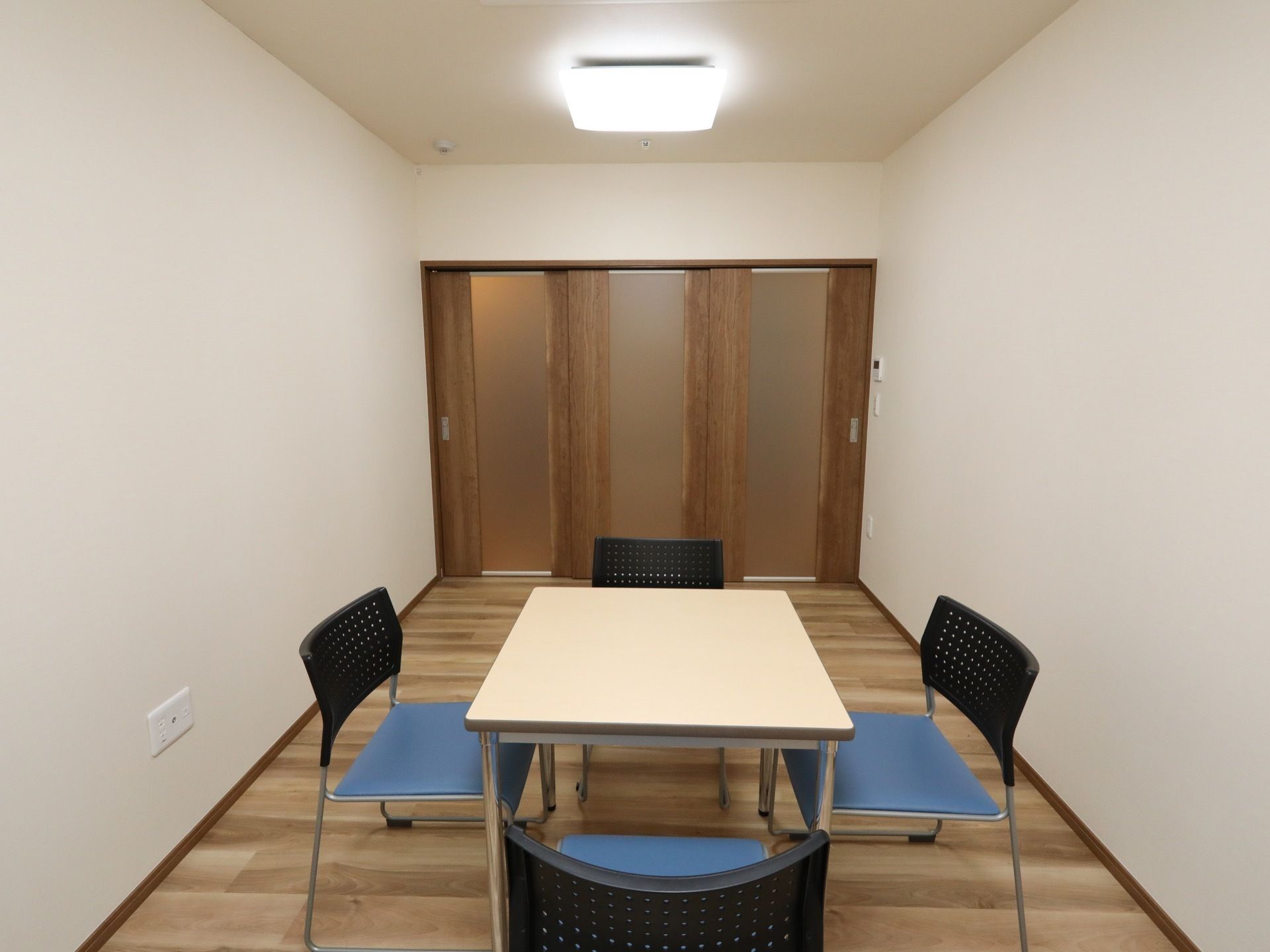 白い壁の部屋の中央にテーブルがあり、その周りに青色の椅子が置かれている。奥には、木製の３枚扉がある。