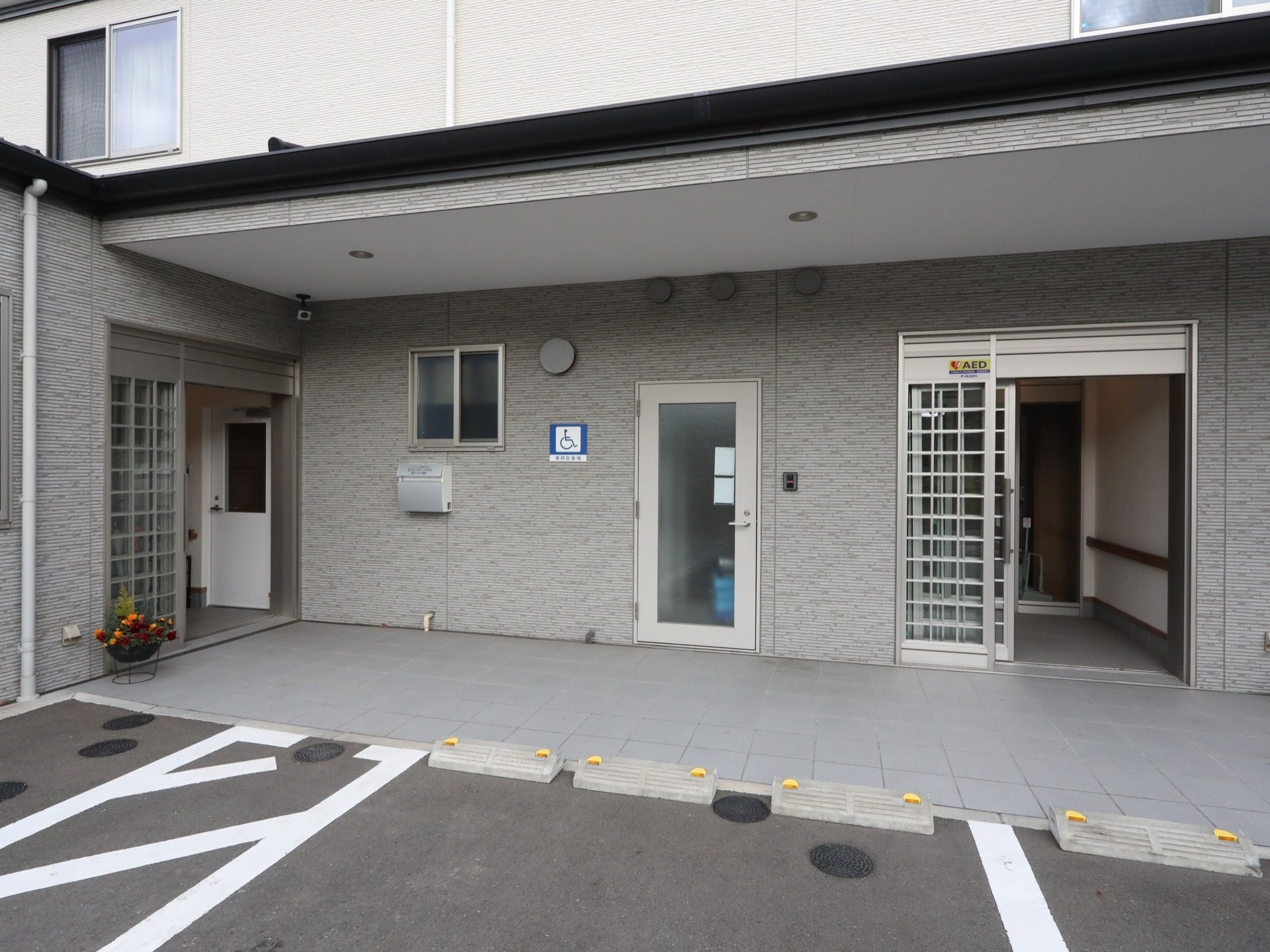 施設の前に駐車スペースがある。車止めが置かれており、白線が引かれている。建物の壁には、車椅子が書かれたプレートが取付けられている。