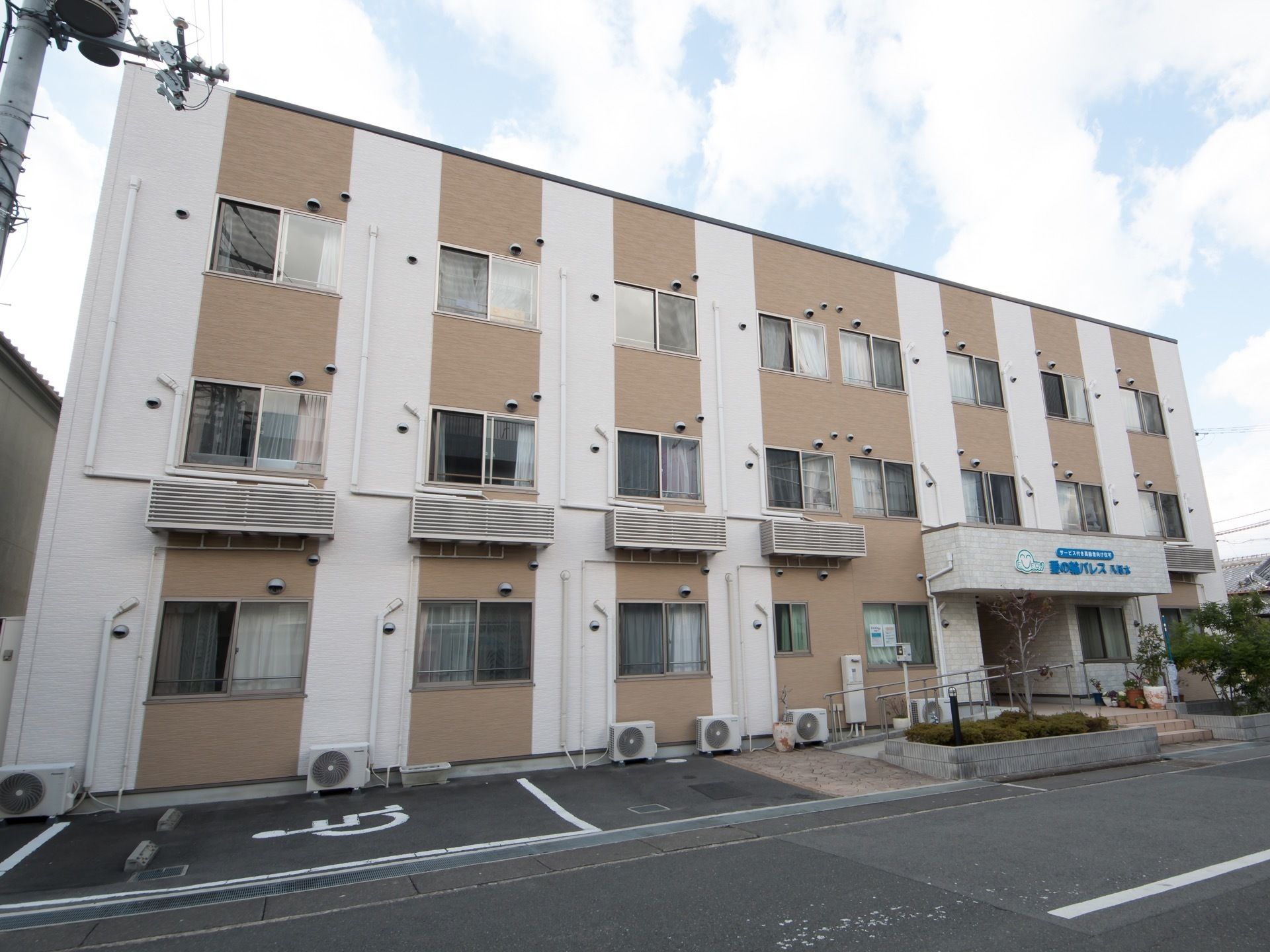 ベージュと白のツートンカラーの３階建て建物。玄関部分はタイルで囲まれており、スロープと手すりが設置されている。