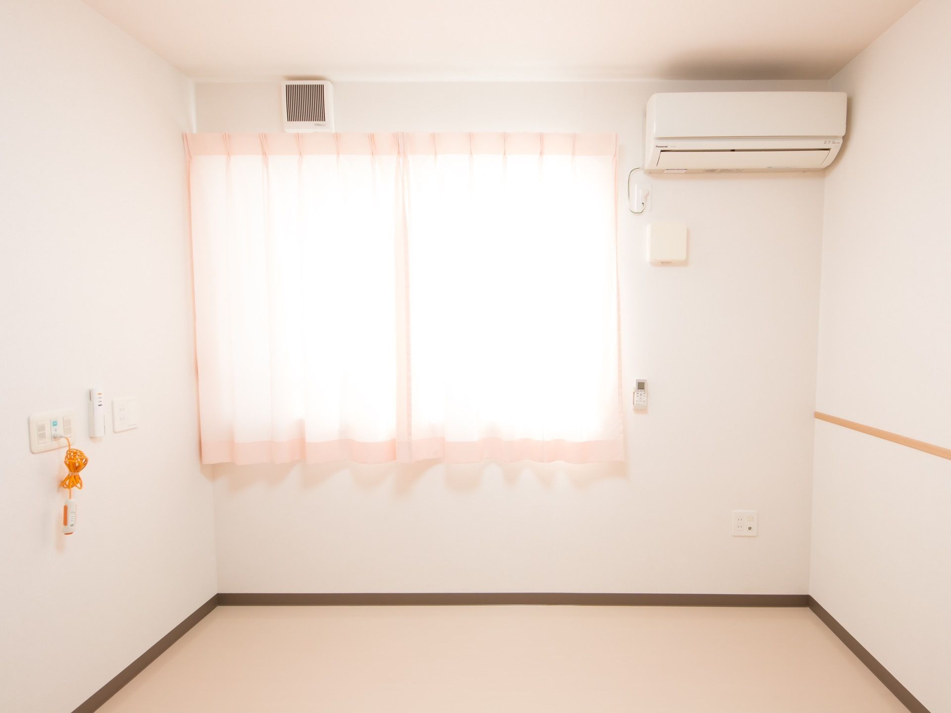 白い壁とベージュの床の明るい居室がある。窓にはカーテンが掛けられており、日当たりが良い。壁にはエアコンが設置されている。
