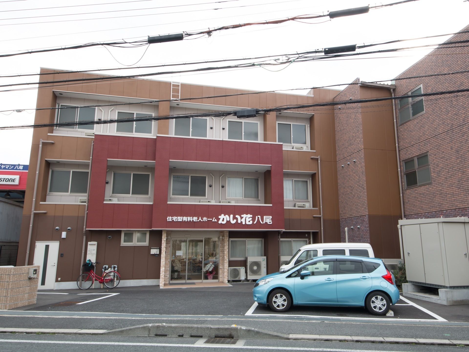 玄関上の外壁に大きく施設名が書かれている。アスファルトで舗装された駐車場の端のスペースに、物置が設置されている。