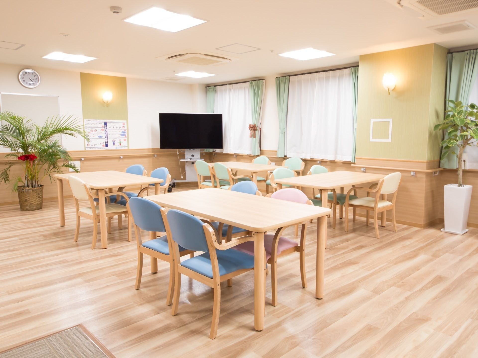 フローリングの床の上に、複数組のテーブルセットが置かれている。壁には手すりが取りつけられており、角には大きなテレビが設置されている。
