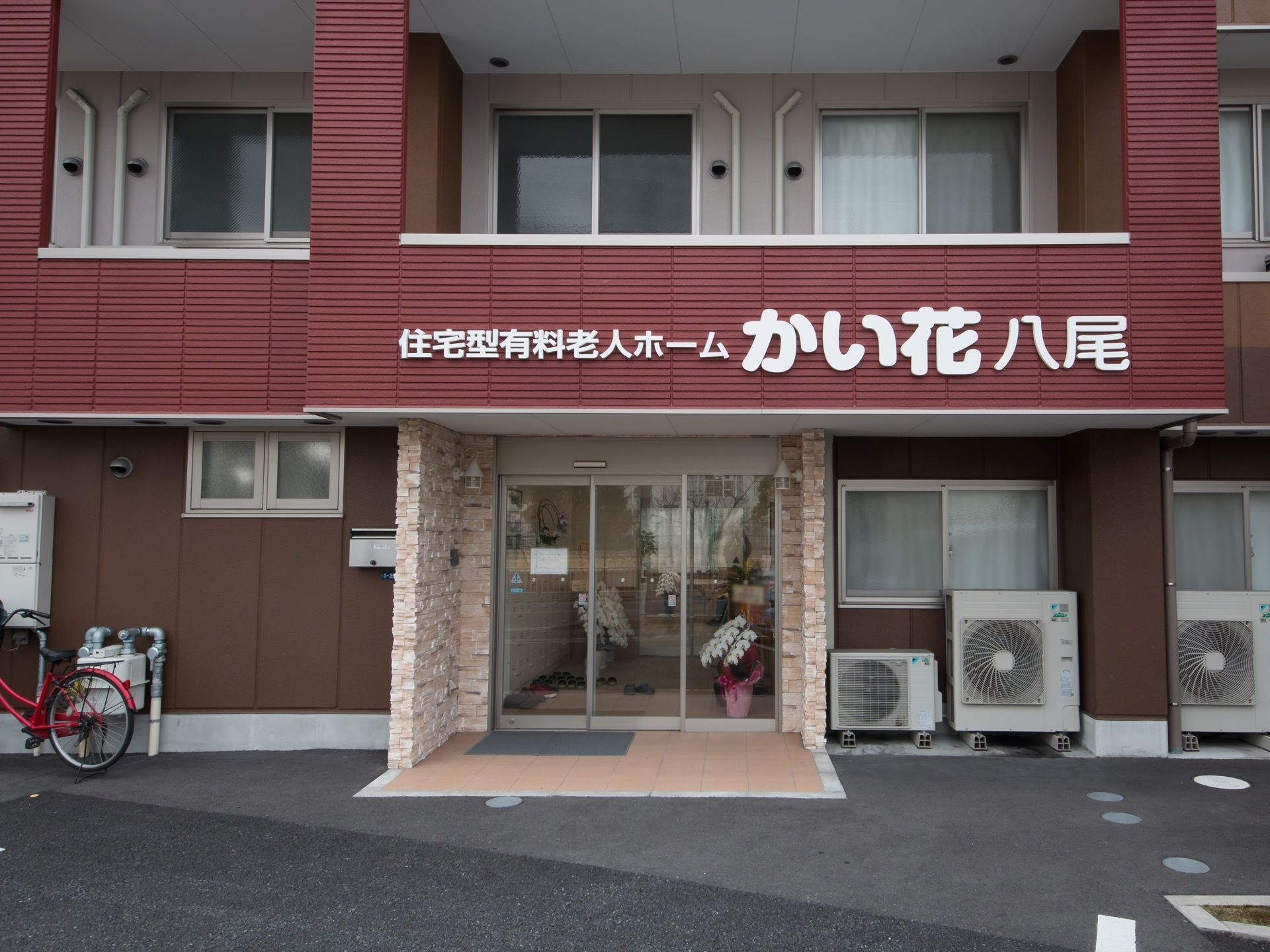 デザイン性のあるお洒落なグループホームで、手前に赤い自転車が１台停められている。駐車場から館内までのアプローチには段差がない。