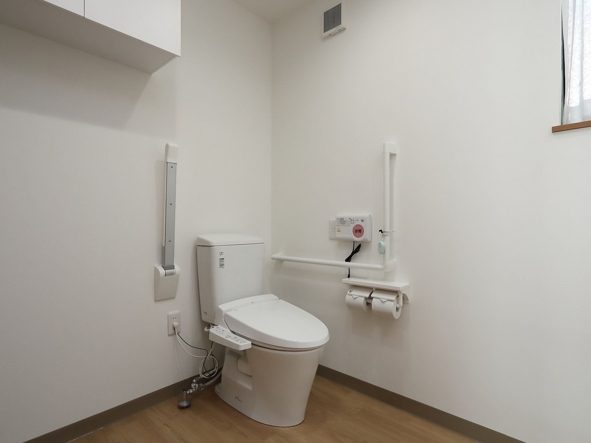 清潔感ある、白色のトイレが備えつけられている。Ｌ字型、可動式の手すりを完備。トイレリモコンが壁に設置されている。