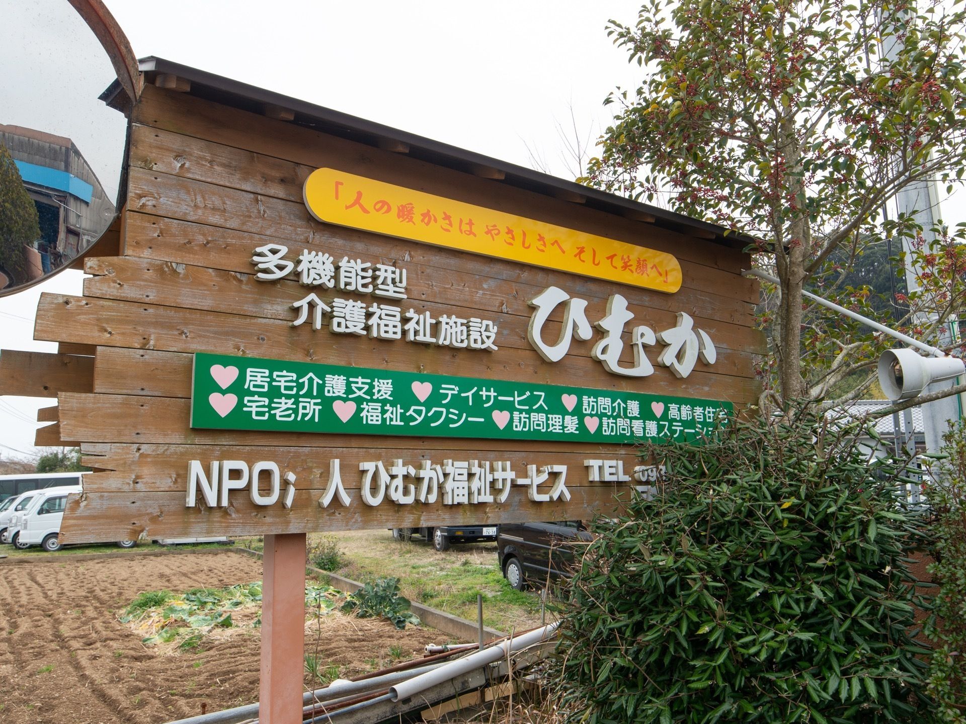 おしゃれな木の板に、大きな文字で施設名が書かれている。また、施設の周りは木や畑などがあり、緑豊かである。