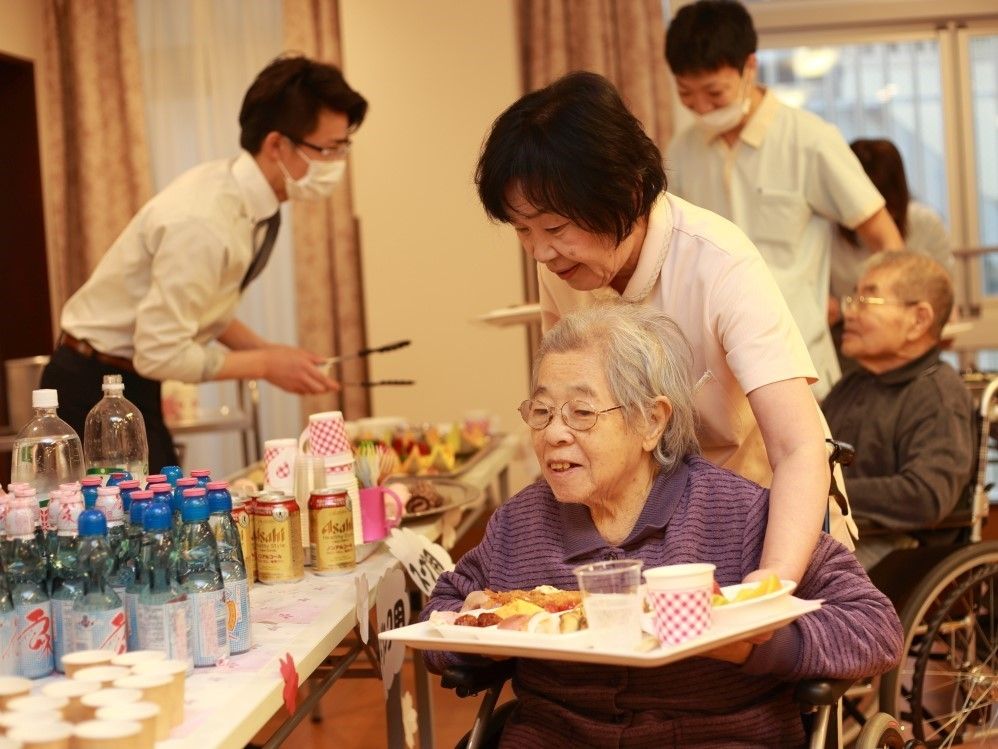 車椅子の入居者が、長机に置かれた飲み物類を選んでいる。