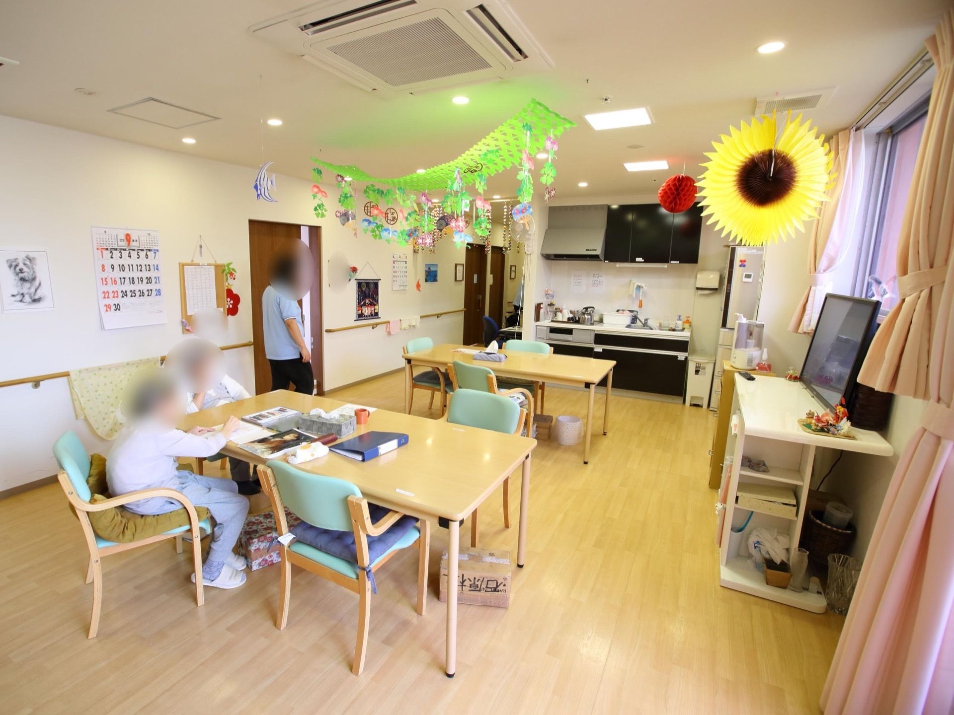 食堂の椅子に入居者様２名が座っている。女性スタッフが１名、テーブルの近くに立っている。窓際にテレビがある。