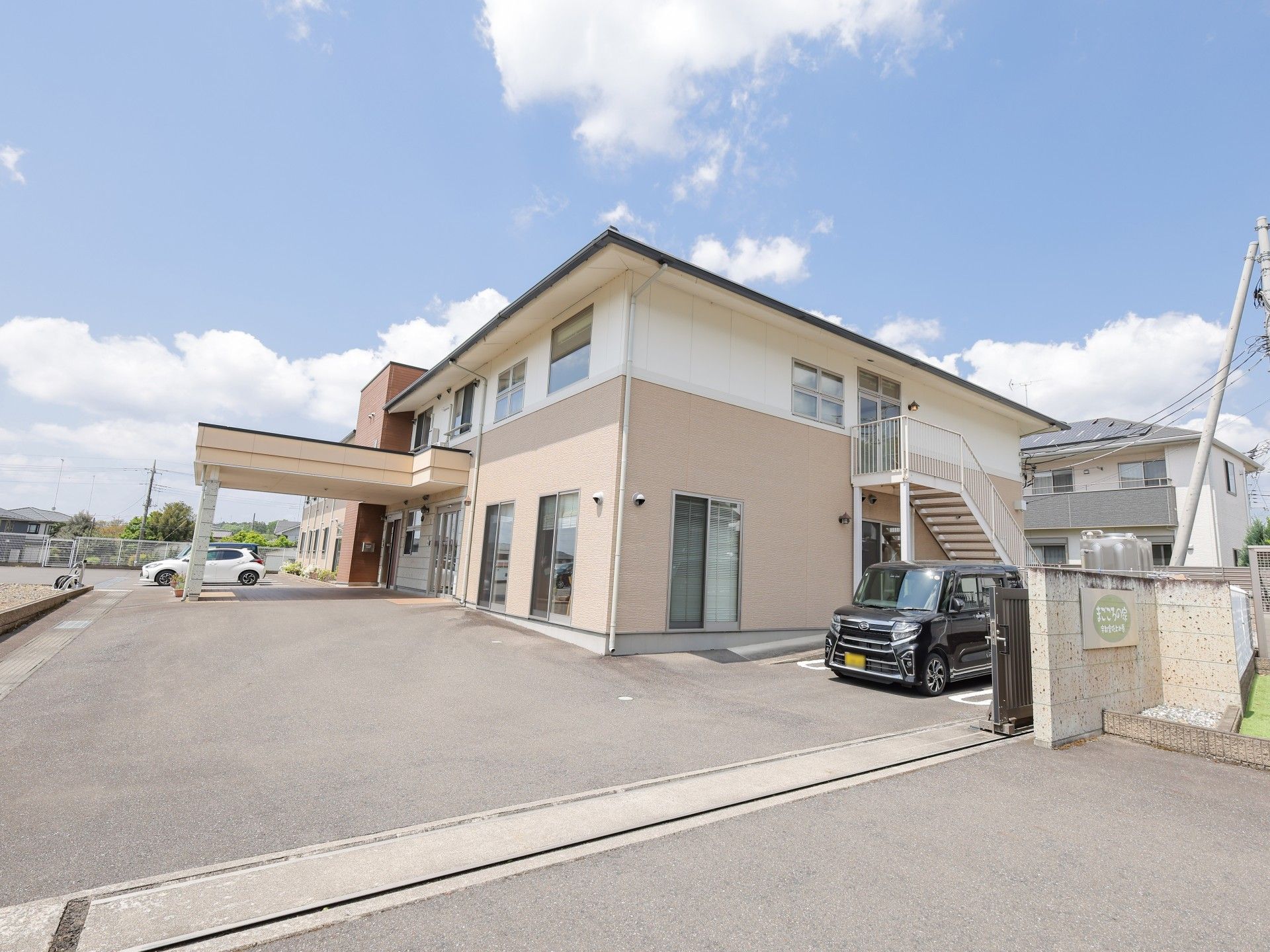 白とベージュの2階建ての建物。玄関には車寄せがあり、その向こうには駐車スペースがある。