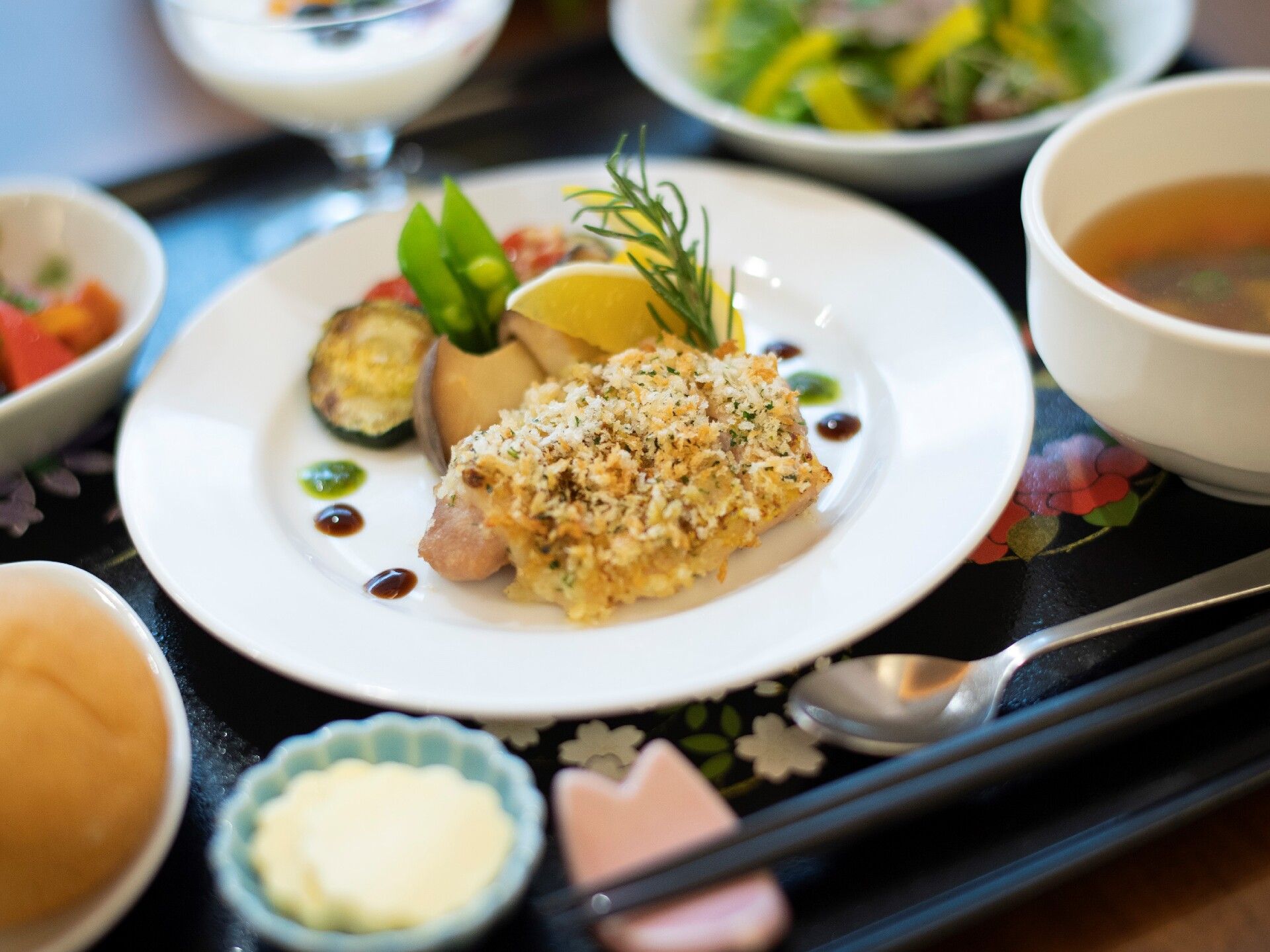 ローズマリーの枝が添えられた香草料理にパン、スープ、野菜料理などが器に盛られ、花柄の黒いトレーに並べられている。