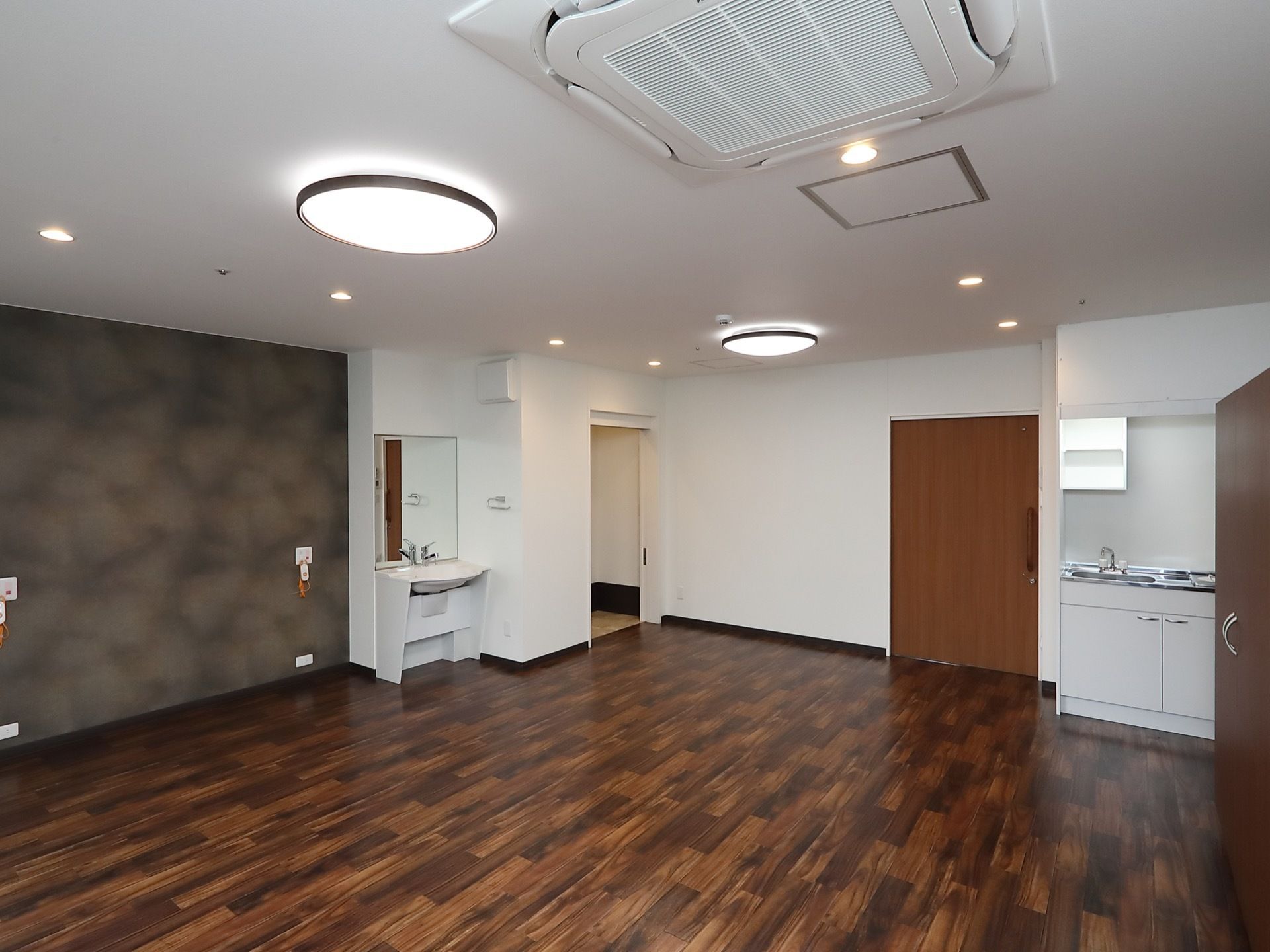 広々とした開放的な空間に洗面台とキッチンが用意されている。床はフローリングになっている。照明と空調も完備されている。