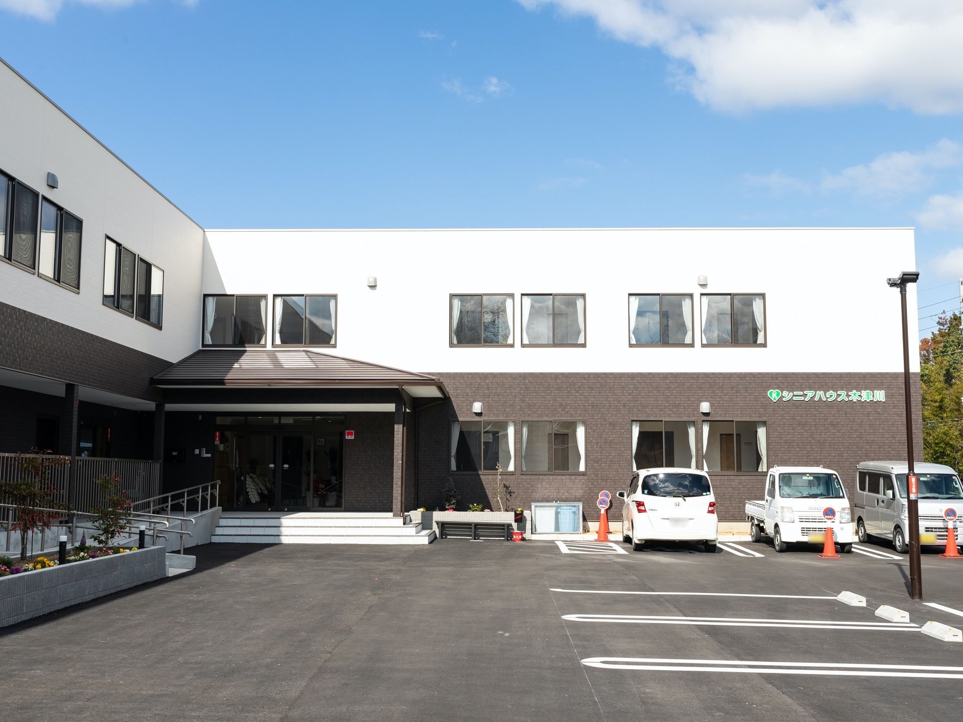 白と茶色の外壁が特徴的である。建物の前には駐車場があり、普通車やトラックが停められている。玄関前に階段とスロープが設けられている。