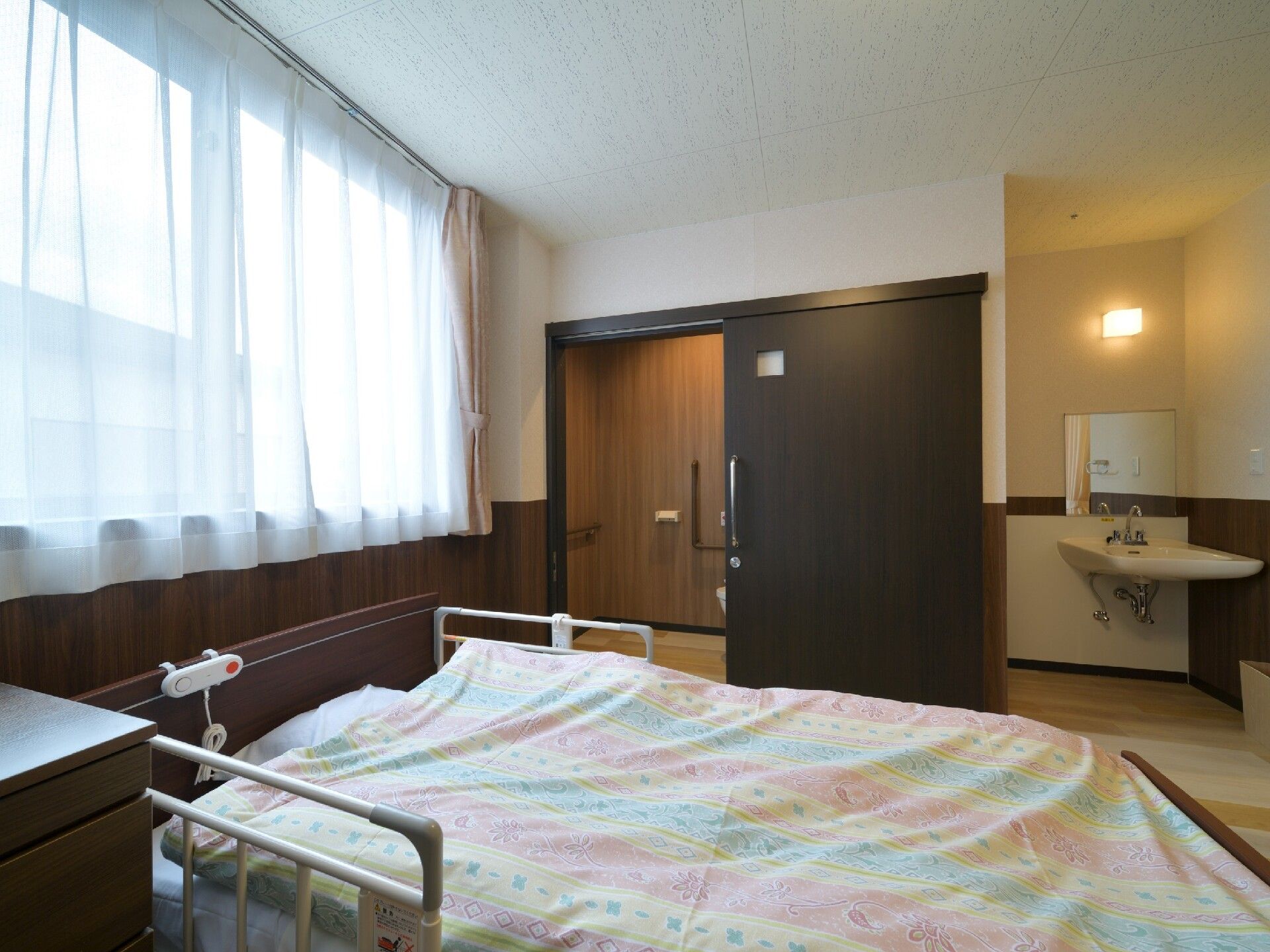 ベッドに寝ている高齢者の女性に話しかけている女性スタッフはエプロン姿。微笑みあっている。