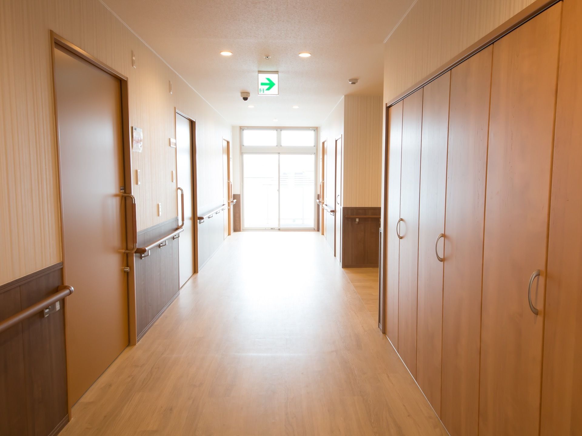 木目仕様のあたたかみのある廊下の様子。壁沿いには滑りにくくつかみやすい手すりを設置している。天井には非常用出口の方向が大きく記されている。