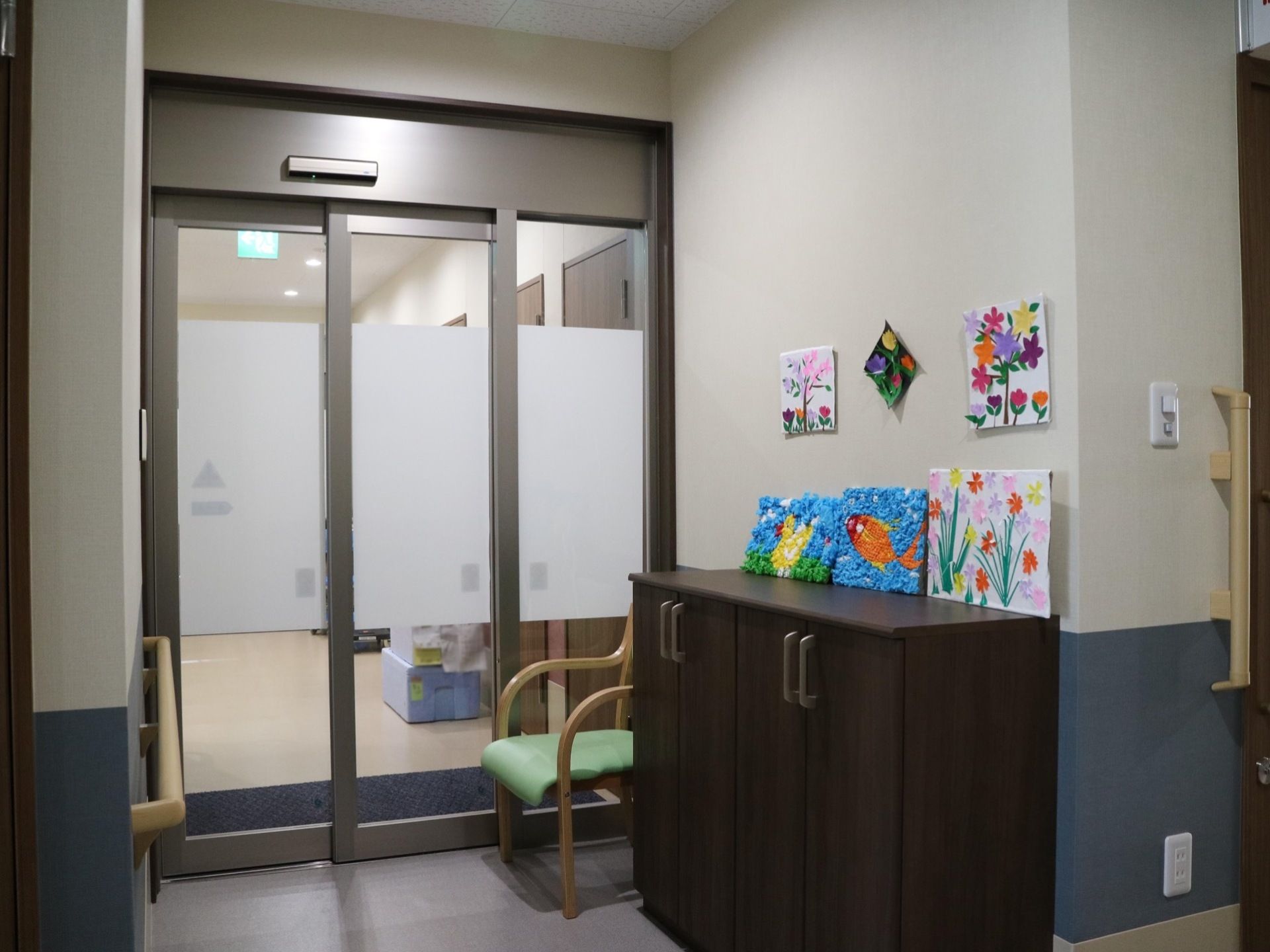 廊下の途中にガラス張りの自動ドアが設置されている。手前にはひじ掛け付きの椅子や、扉付きの棚が置かれている。
