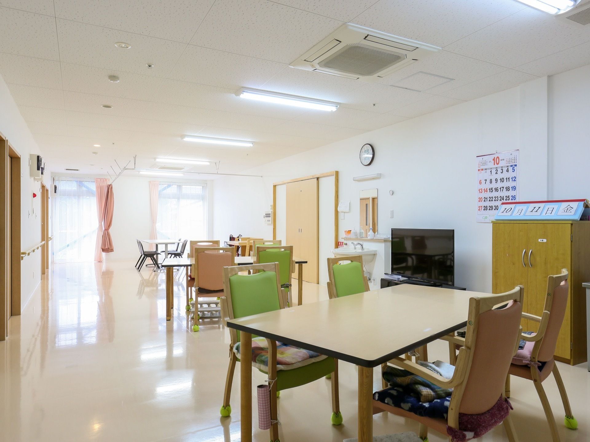 共有スペースにはテーブルと椅子がある。壁には時計、カレンダーが掛けられている。洗面台の横にはテレビが置かれている。