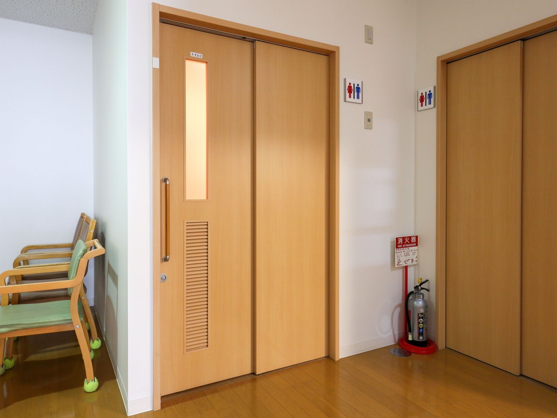 トイレの個室が2つある。扉は茶色で、扉横にトイレマークのプレートが付けられている。消火器も置かれている。