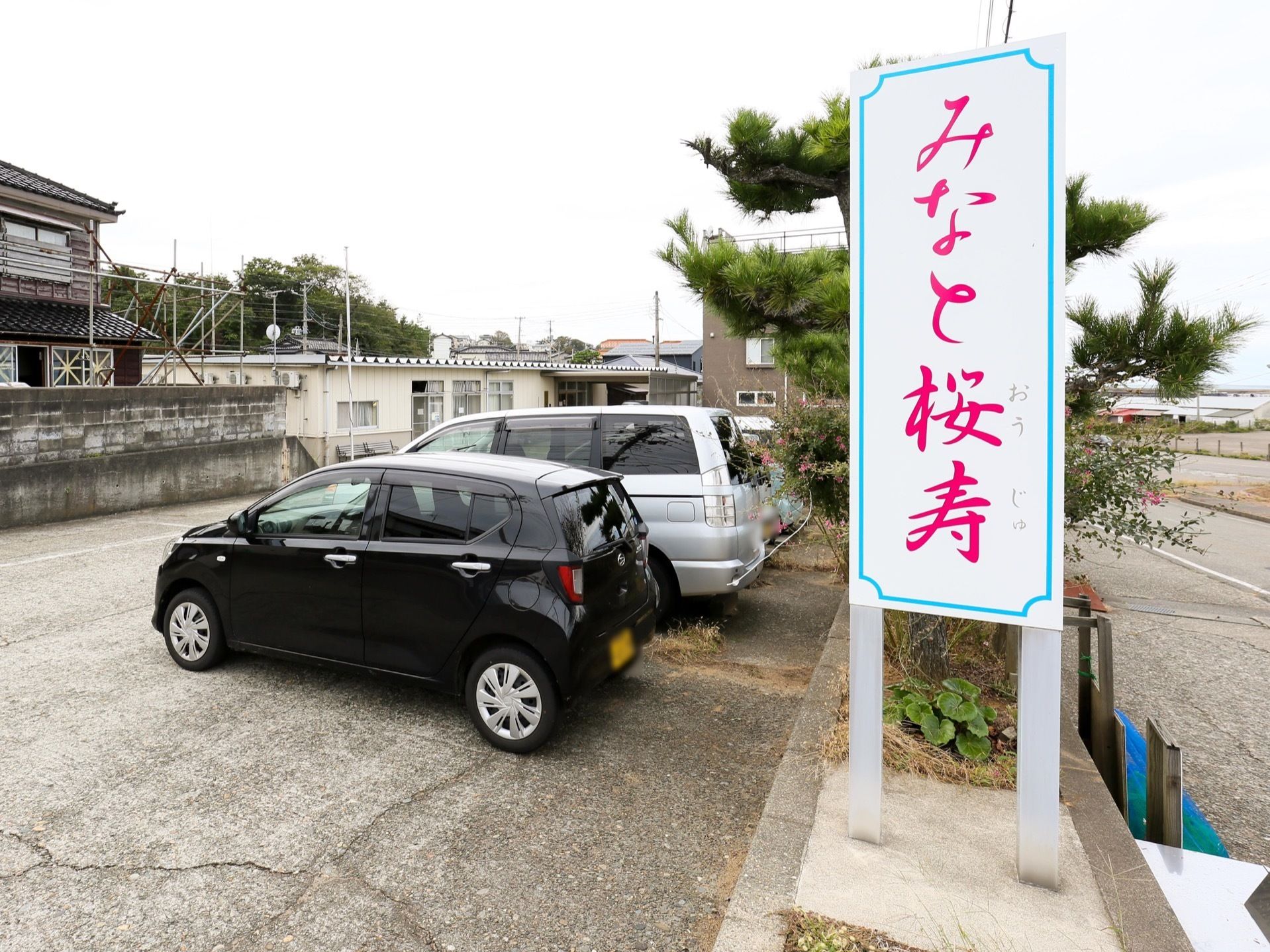 施設には縦型の看板がある。水色の枠の中にピンク色で施設名が書かれている。駐車場には4台ほどの車が止められている。