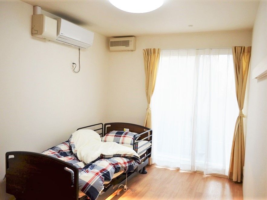 エアコンが設置されたフローリングの部屋にはカーテン付きの窓があり、窓辺にはベッドが置かれている。