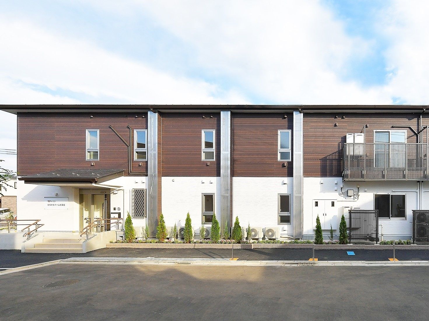横に長い２階建ての建物は白とブラウンで、玄関先には屋根が付き、駐車場もある。
