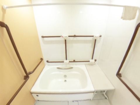 一面だけ木目調の模様が付いた白い浴室には四角い浴阻喪と手すりが設置され、浴槽の横にはベンチもある。