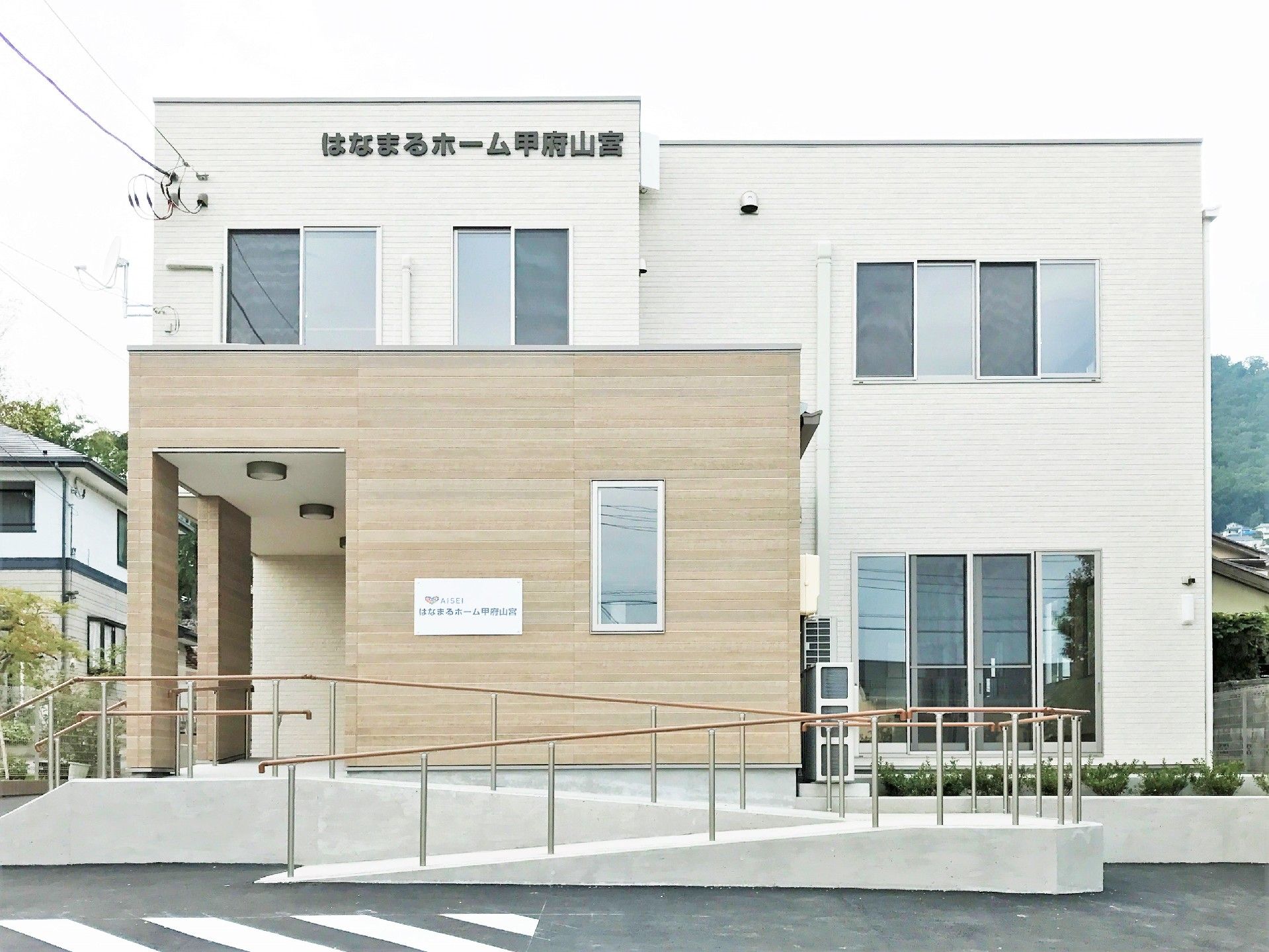 白を基調とした2階建ての建物の周りには自然が多く、玄関先には手すり付きのスロープが付いている。