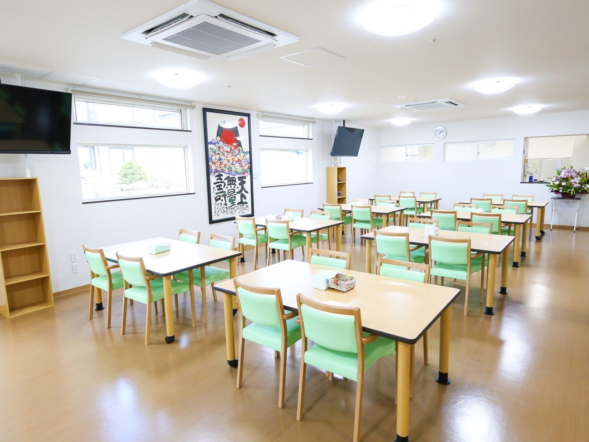 テーブル席がたくさん設置された食堂で、それぞれのテーブルの上にティッシュ箱が置かれている。吊り下げ式のテレビが２台ある。