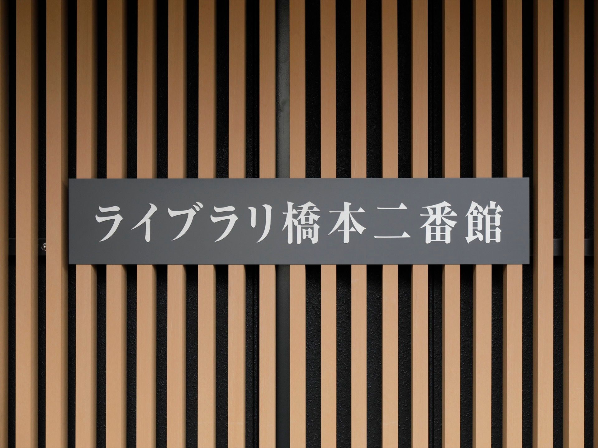 黒い壁に茶色の木材が格子状に貼られた外壁に施設名が書かれた看板が取り付けられている。黒い板に白字で書かれている。