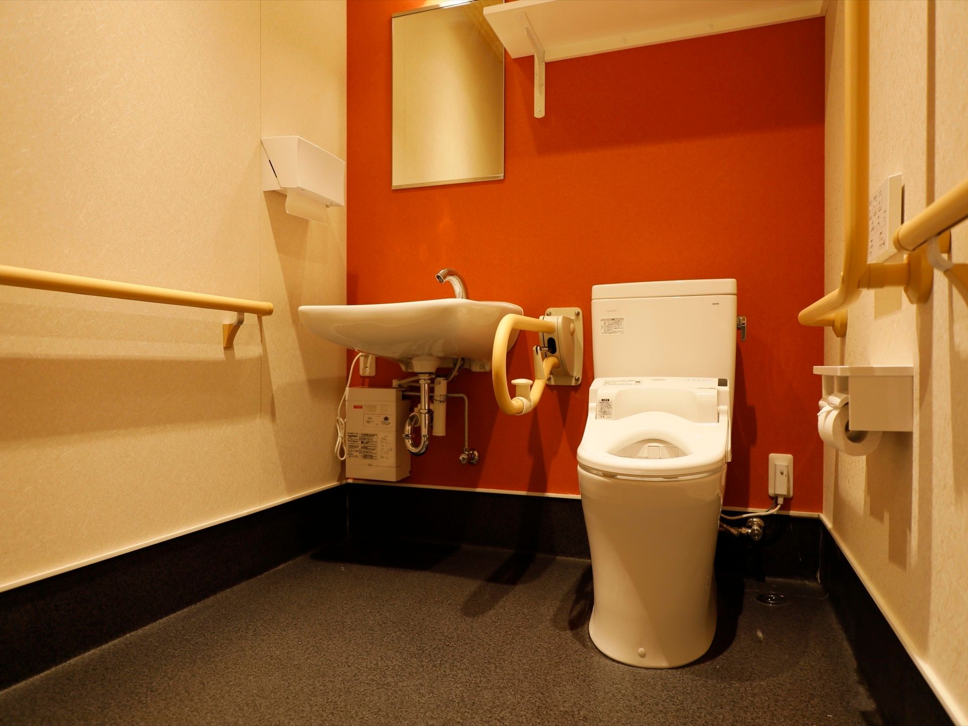 黒い床に赤と白の壁のトイレ個室である。温水洗浄便座を取り付けた白い便座があり、その隣に流し台が設置されている。