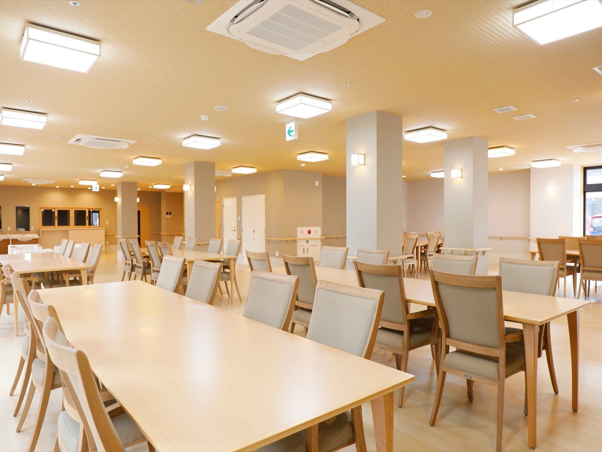 とても広いダイニングに８人で利用できる長いテーブルと椅子がたくさん並べられている。奥にはガラス窓が閉められたキッチンがある。