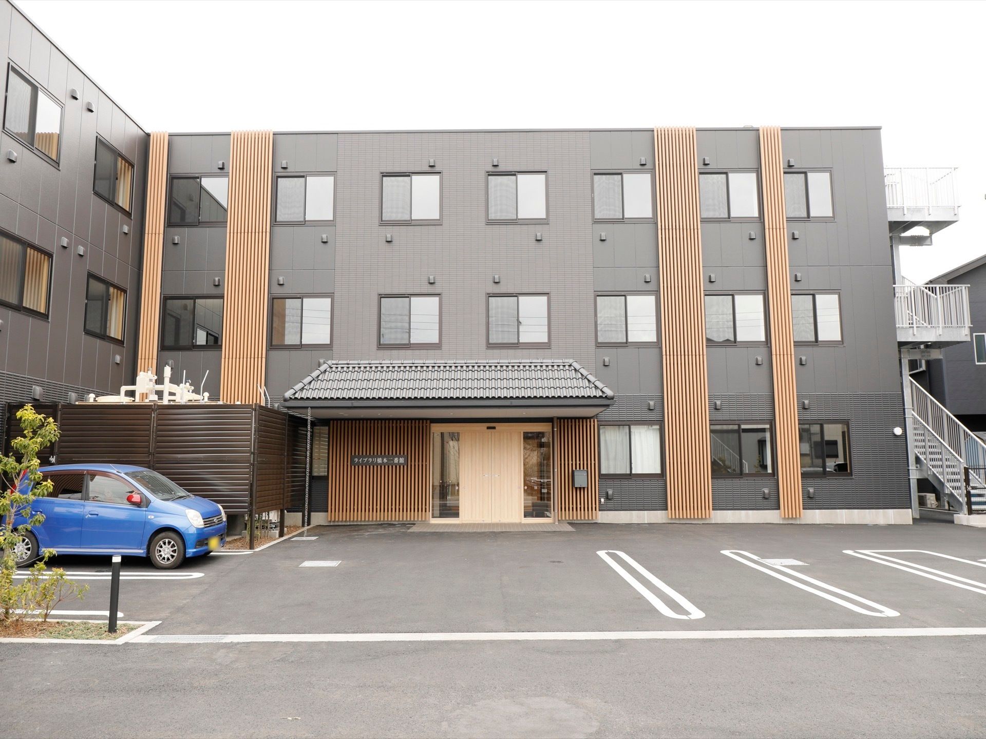 グレーの外観で木製の柱が縦にたくさん並べられている。建物の前は駐車スペースになっており、青い自動車が１台停まっている。