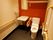 赤と白の壁の個室トイレがある。便座の横の壁に折り畳み式の手すりがある。天井近くには棚が取り付けられている。