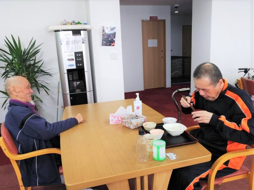 二人の男性入居者がテーブルにつき、一人は食事を食べていて、もう一人はそれを眺めている。