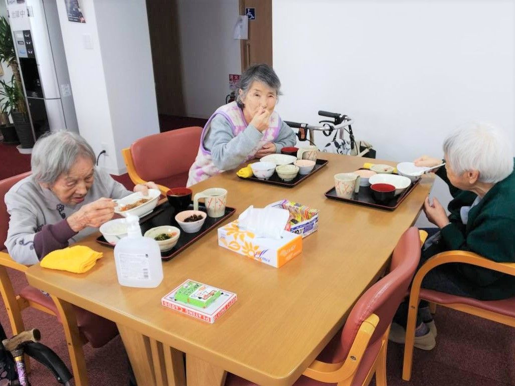 3名の女性入居者が食卓を囲んで食事をしている風景。小鉢が多く、お茶や汁椀などもトレーの上にある。