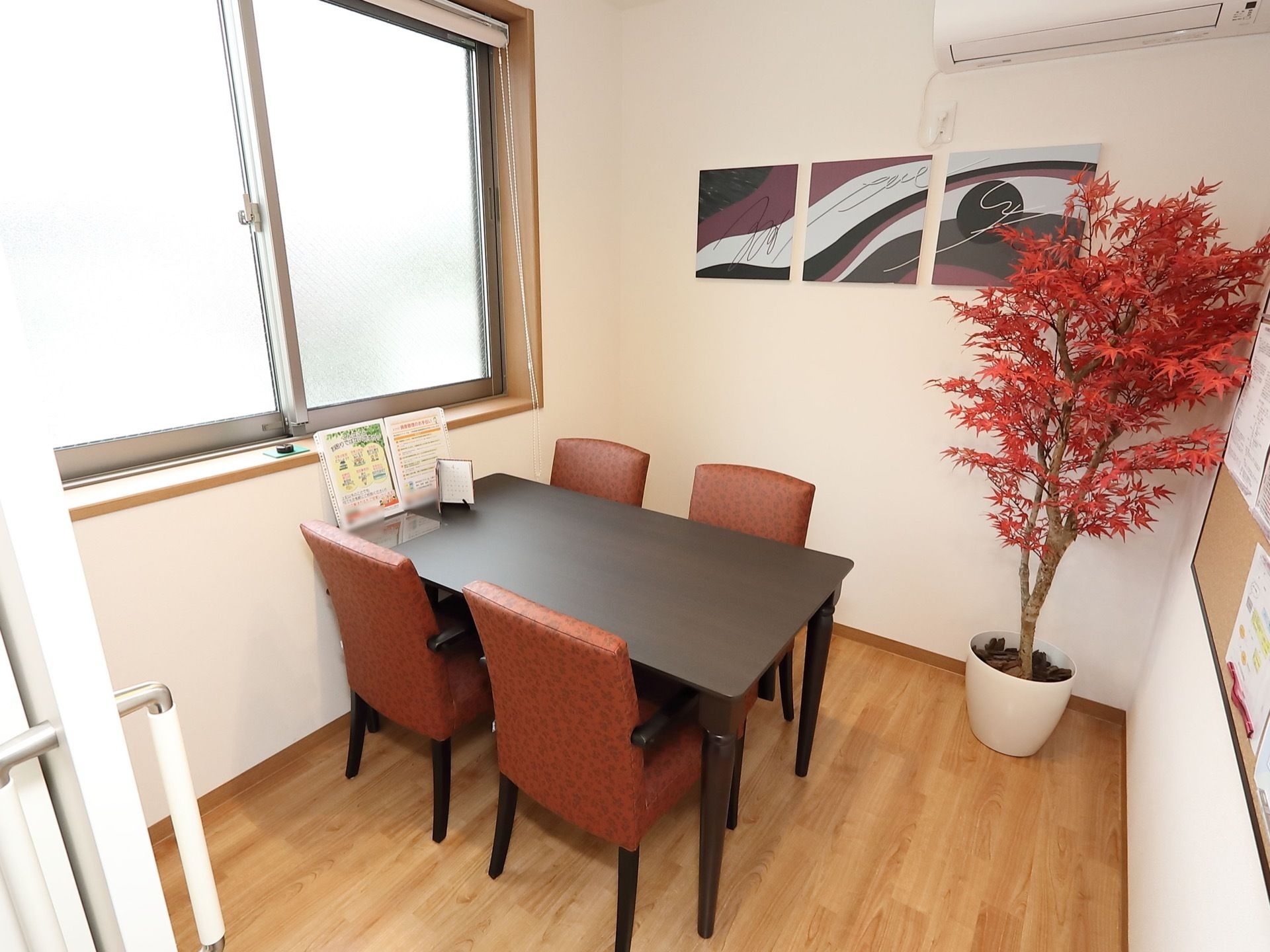 紅葉の鉢植えが飾られている部屋に、テーブルと椅子４脚が設置されている。３枚組の平面作品や掲示板が壁に取り付けてある。