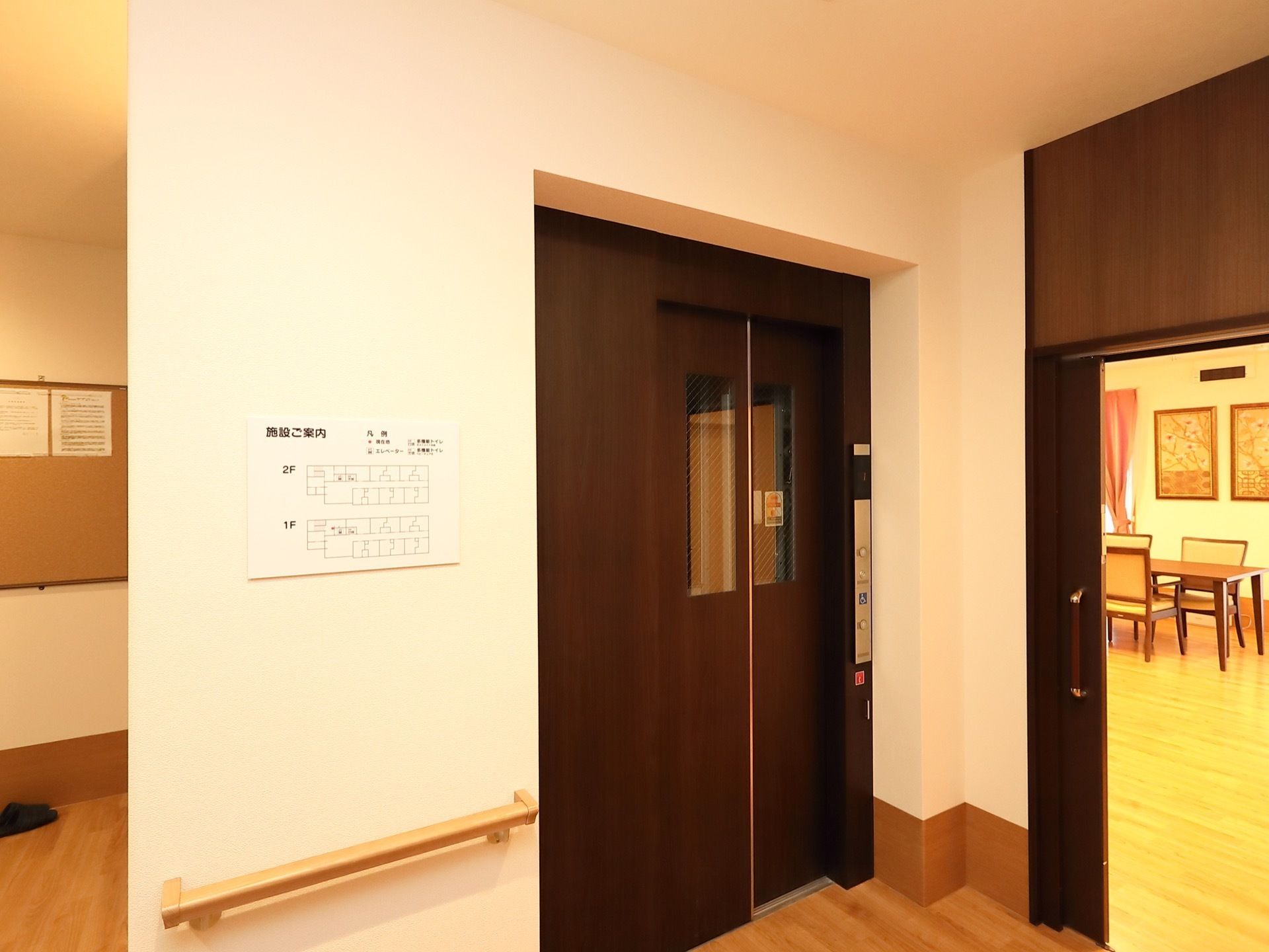 エレベーターが設置されており、左側に施設の案内図が掲示されている。右側の引き戸の奥に、テーブルや椅子が置いてある。