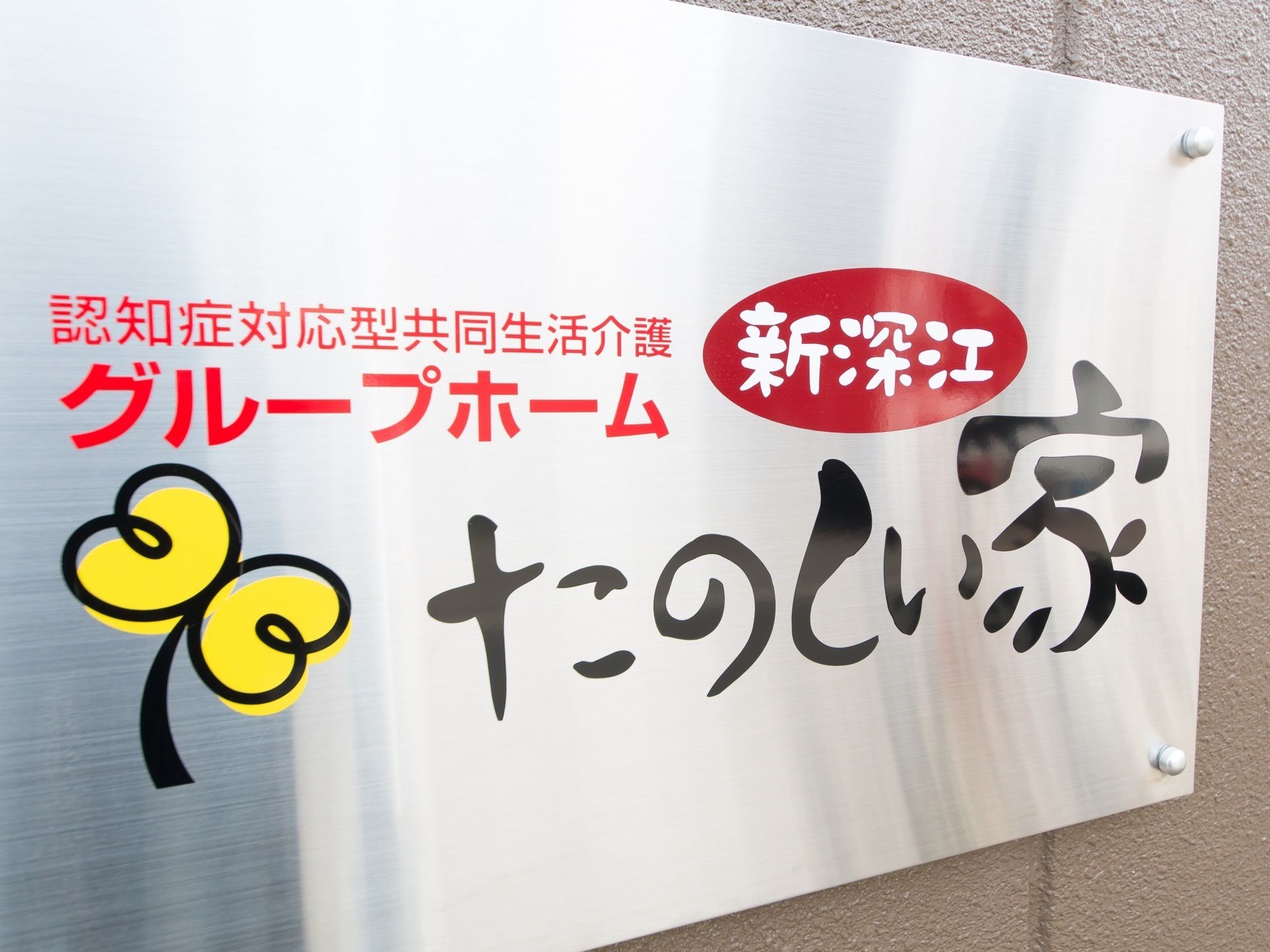 施設名称と提供サービス形態が書かれた銀色の看板が設置されている。２枚の葉っぱをイメージしたマークも添えられている。
