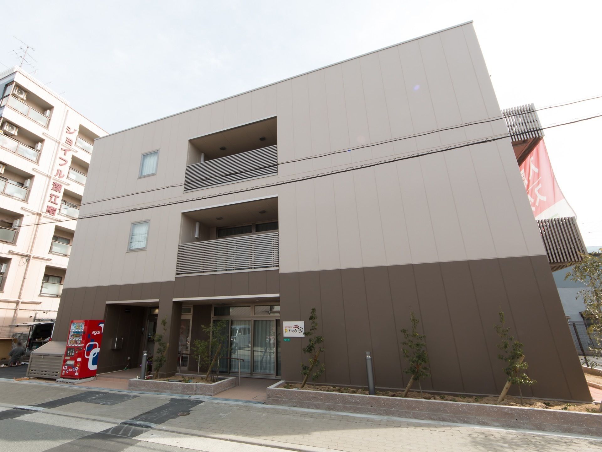 ツートンカラーの外壁になっている３階建ての施設である。玄関はひさしの奥にある。歩道に面した部分に花壇がある。