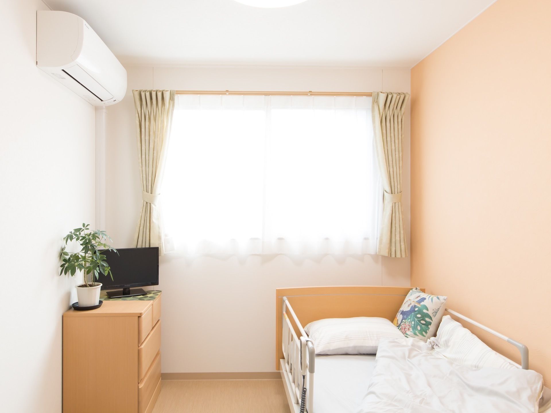 居室には大きな窓がある。転落防止柵つきのベッドやエアコンを完備している。衣類収納棚の上にテレビと観葉植物が置かれている。