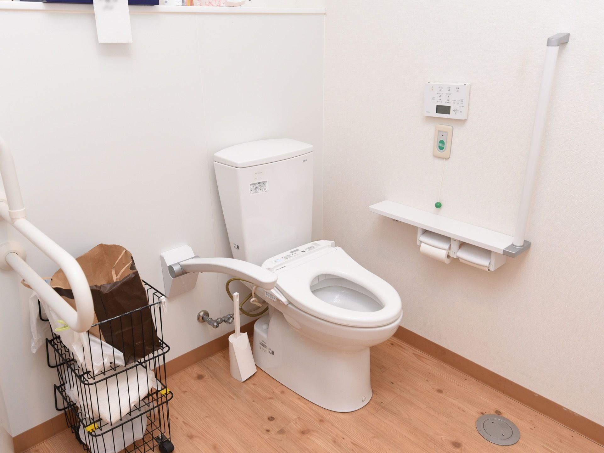 広い個室トイレである。床はあたたかみのある木目調で、壁は白く清潔感がある。手すり、非常ボタンが設置されている。