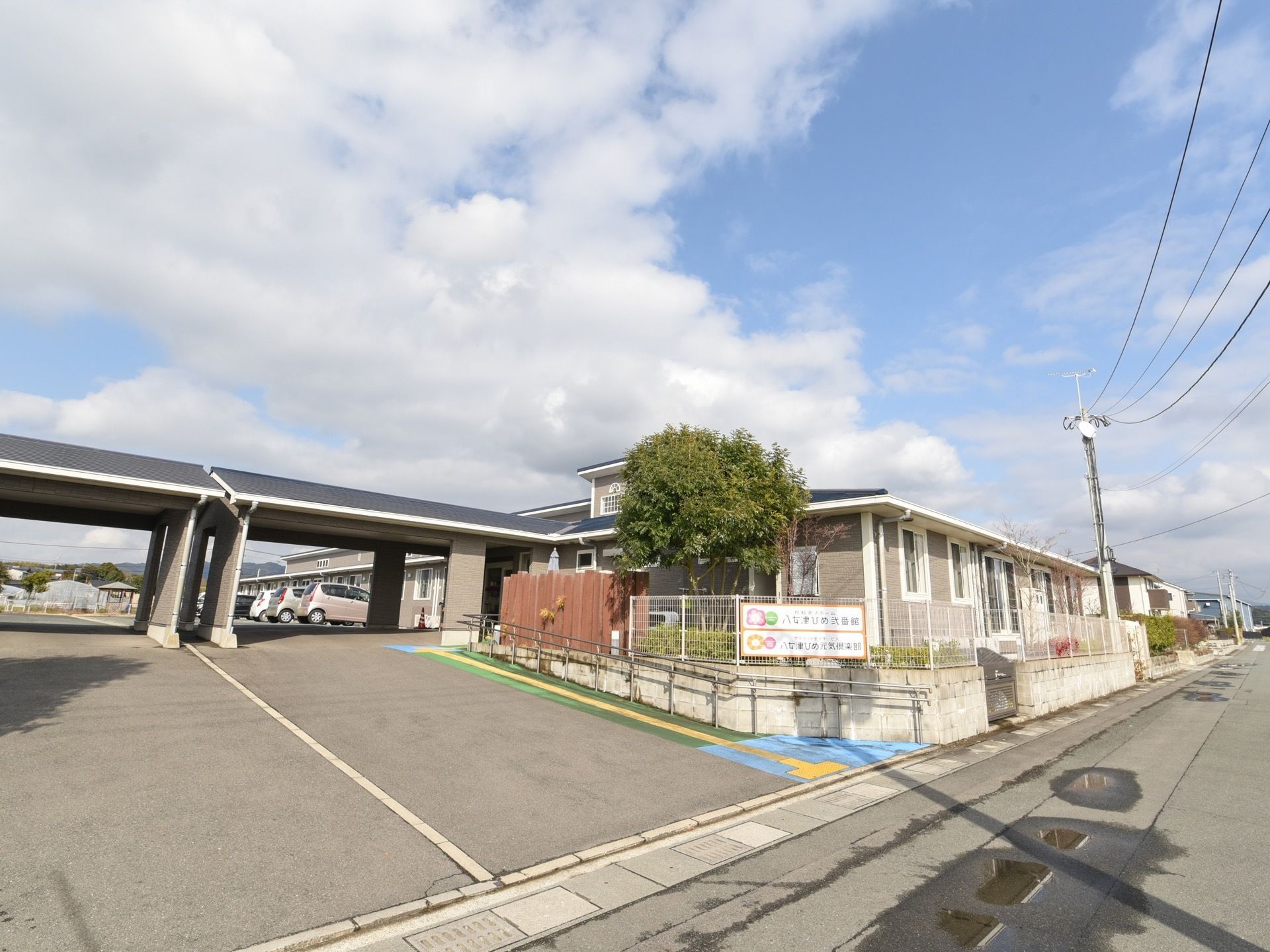 施設の外観である。駐車スペースの入口に、施設名の看板を掲示している。駐車スペースから玄関へと続く屋根がある。