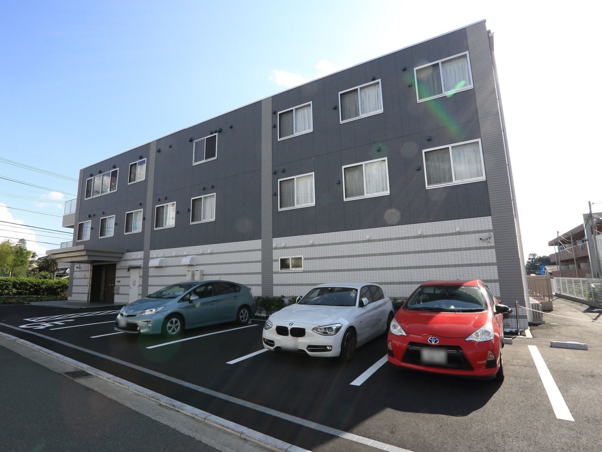 晴天の日の3階建施設の外観である。建物の左側に施設入口を設け、外壁に沿って駐車スペースを確保している。