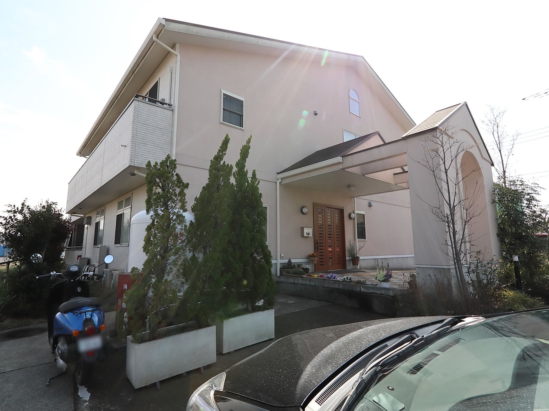 一軒家の住宅のような外観の建物である。建物の周りには植え込みや庭があり、正面には駐車スペースが設けられ、バイクや自動車が停められている。