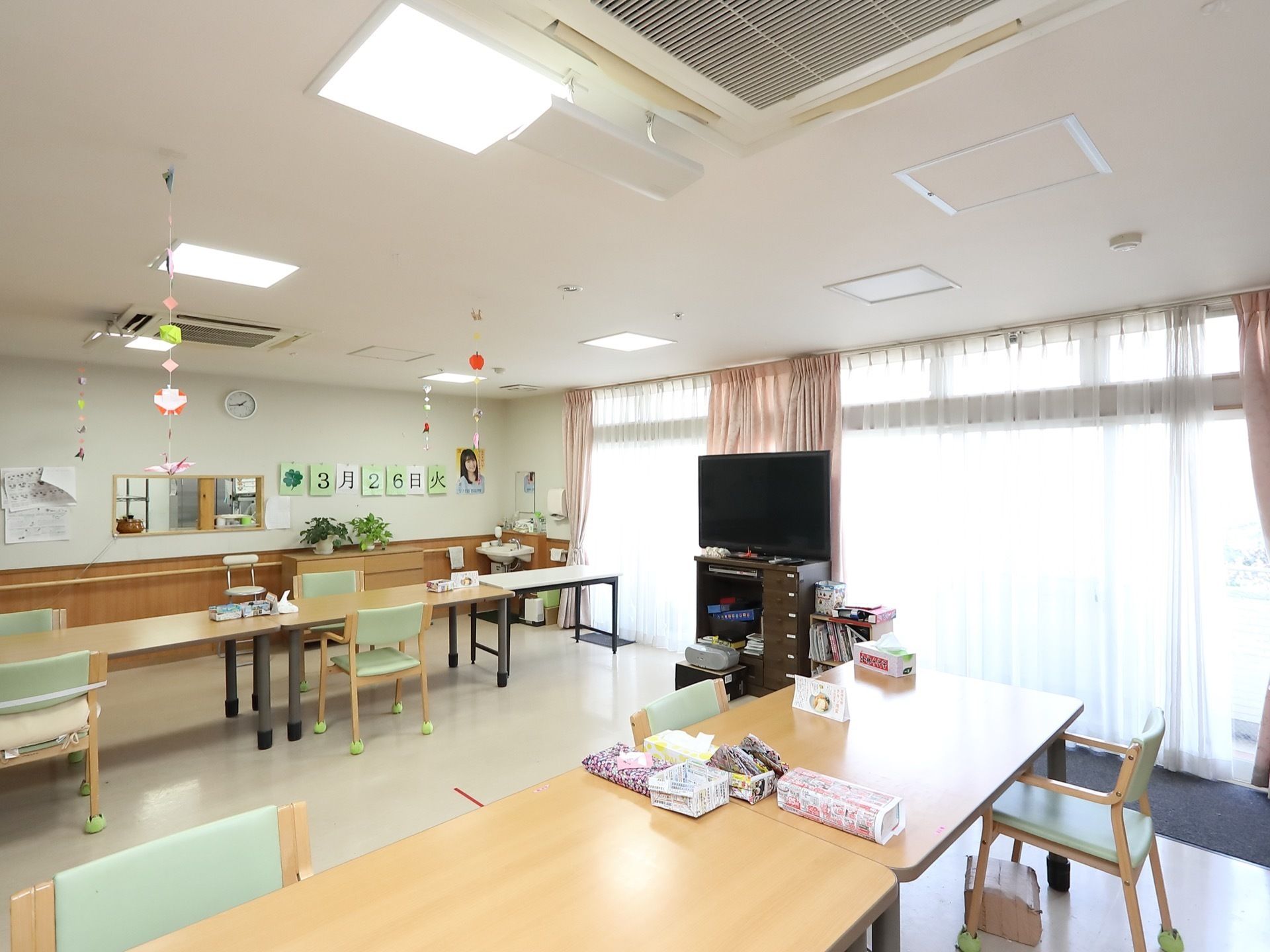広く開放的な共有スペースである。食堂兼用で奥にはキッチンが見える。大きな窓からはカーテン越しに外光が差し込み、とても明るい。