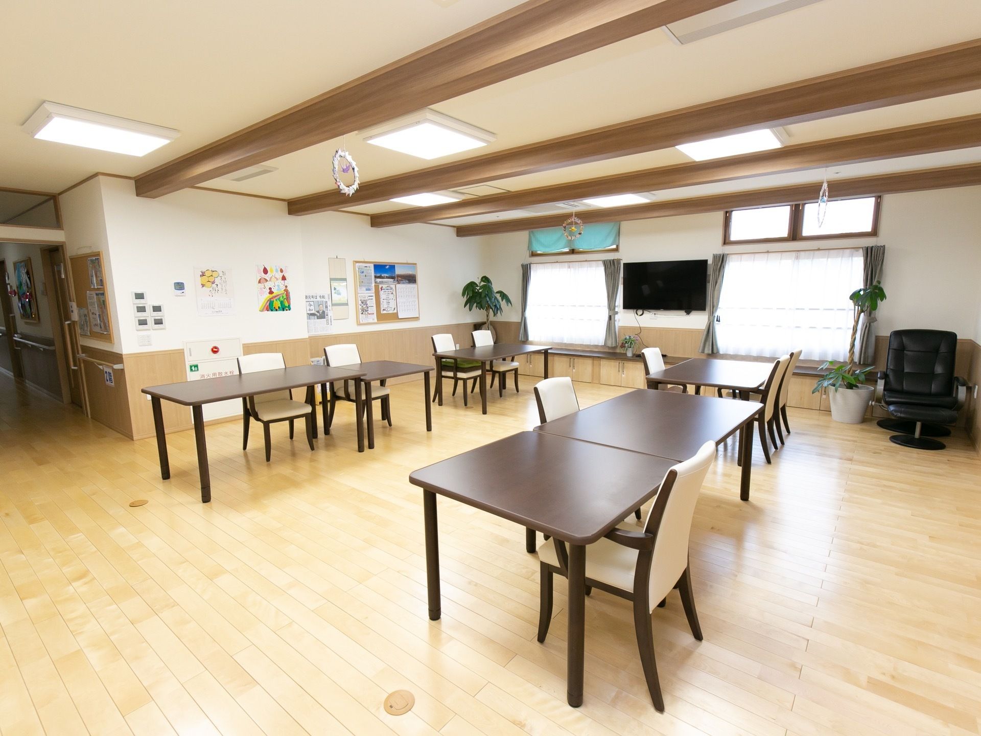 梁見せ天井のリビングに、テーブルと椅子が複数設置されている。窓の側には、観葉植物や足台付きの肘掛け椅子が置いてある。